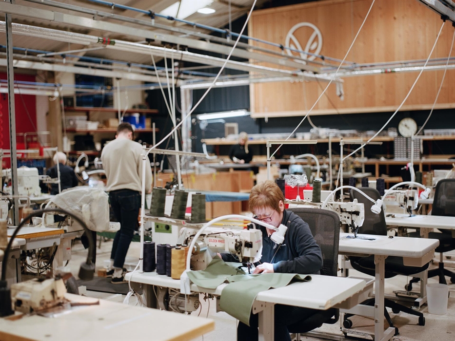 A Grandmaster sews denim at a workstation inside the Hiut Denim factory. Other team members work in the background beneath a wooden wall featuring the
