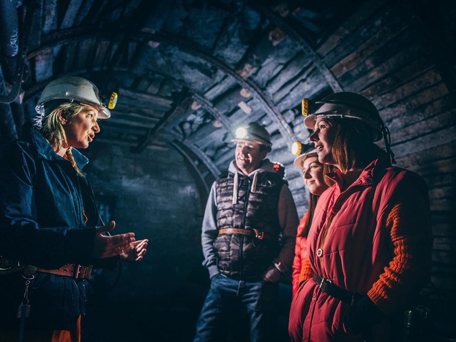 A guide is dressed in dark blue overalls and a white hard hat. She is talking to three visitors, a man, woman and teenage girl. They are also wearing