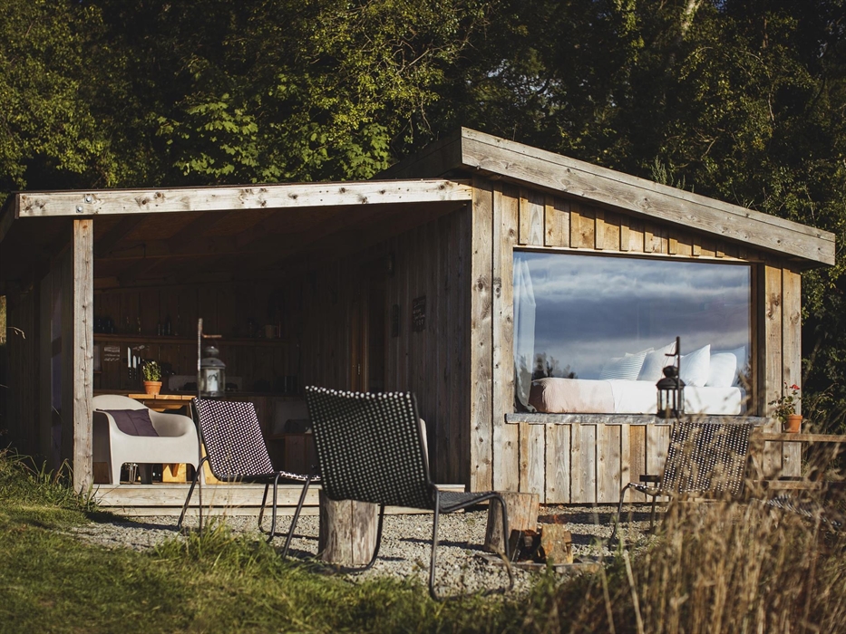 Wood cabin set amongst woodland.  There is a covered outdoor camp kitchen and a firepit to the foreground. Inside is a bed made up with a view out of