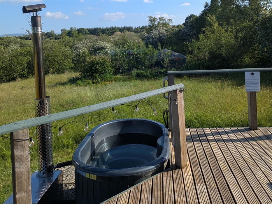 Grey fibreglass wood fired hot tub off wood deck, with chrome safety handles on posts, stove with chimney to left of photo. Wildflower meadow and tree