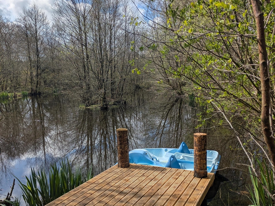 In the foreground is a wood jetty with a pale blue pedalo on the lake behind. There is an island with trees and trees and irises on the lake shore. Th