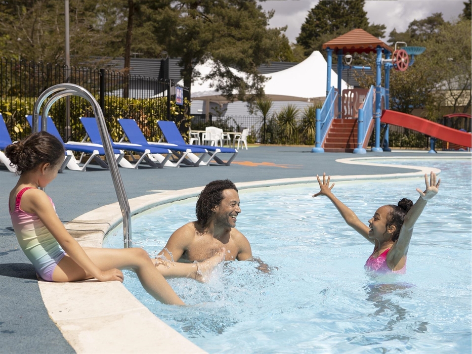 2 girls and a man are playing in an outdoor pool. In the background: a water slide and sun loungers.