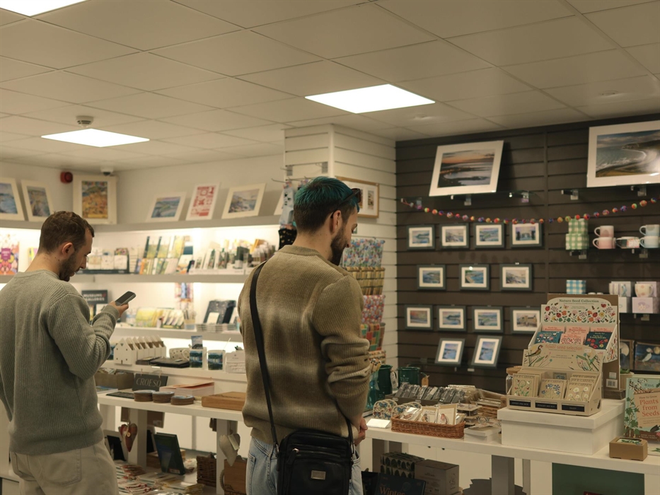 Two men are standing in the shop with a table of a variety of shop items and a wall display in front of them.