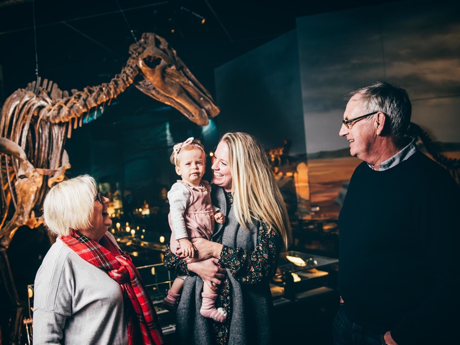 grandparents, mum and small girl. All smiling. Standing in front of a large dinosaur skeleton.