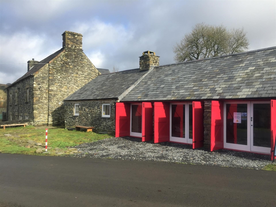 Exhibition exterior showing 3 sets of red shutters with the farmhouse in the background