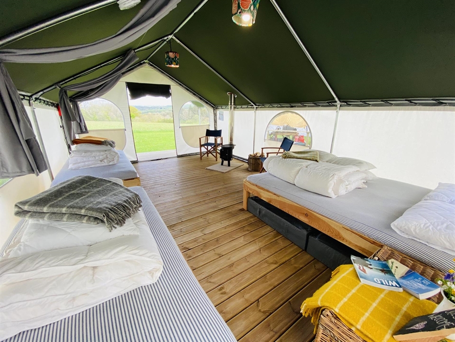 Inside a glamping tent showing a king size and two single beds, with a a log burner and chairs on a decking style floor. Background of green fields