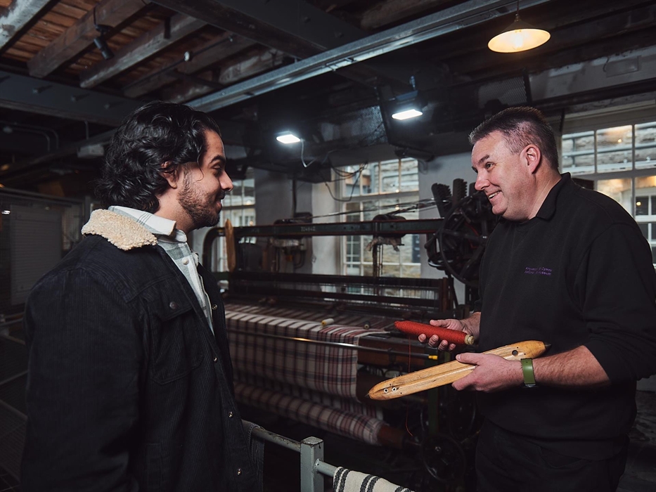 A visitor talks to a craftsman in the weaving workshop. The craftsman explains the weaving process. He is holding a bobbin of red thread and a wooden