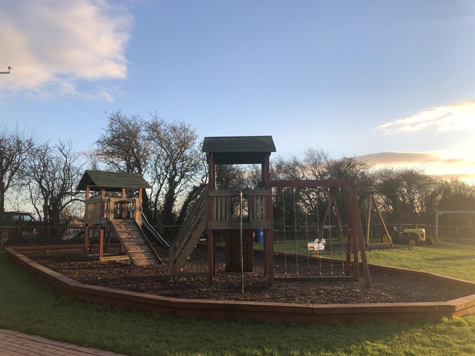 A wooden playground with climbing walls, a fireman's pole, swings, a slide and a mini tractor on grass and in front of trees.