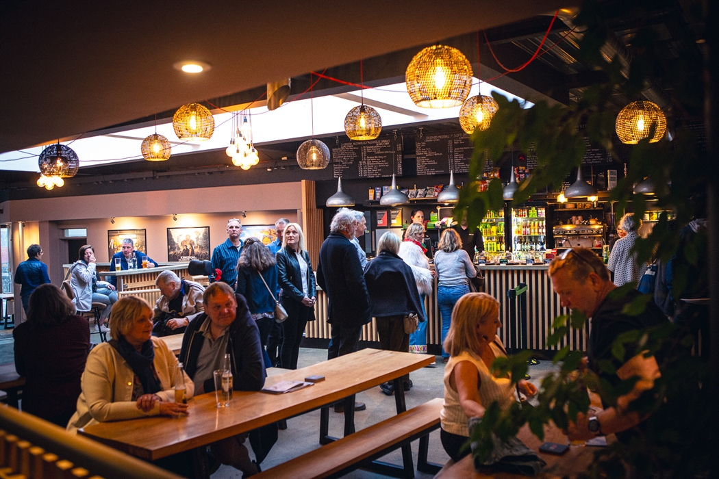 People sit and stand in a busy bar area with wooden tables and hanging lights