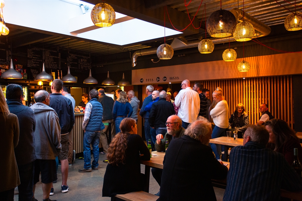 People stand and sit in a warmly light bar area with low hanging lighting and wooden vertical slats towards the rear of the image