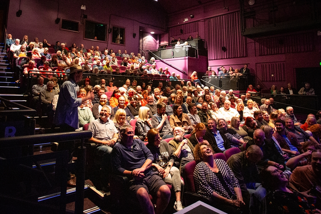 The camera faces an auditorium full of audience members, illuminated by theatre lighting. A man in a denim shirt and hat stands on the steps to the si