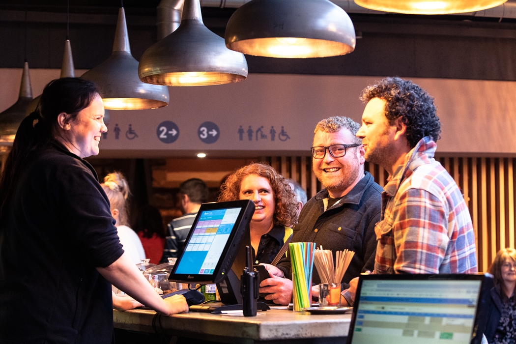 A man with a checked shirt, a man with glasses and a woman with curly hair stand at a bar being served by a lady with a dark ponytail