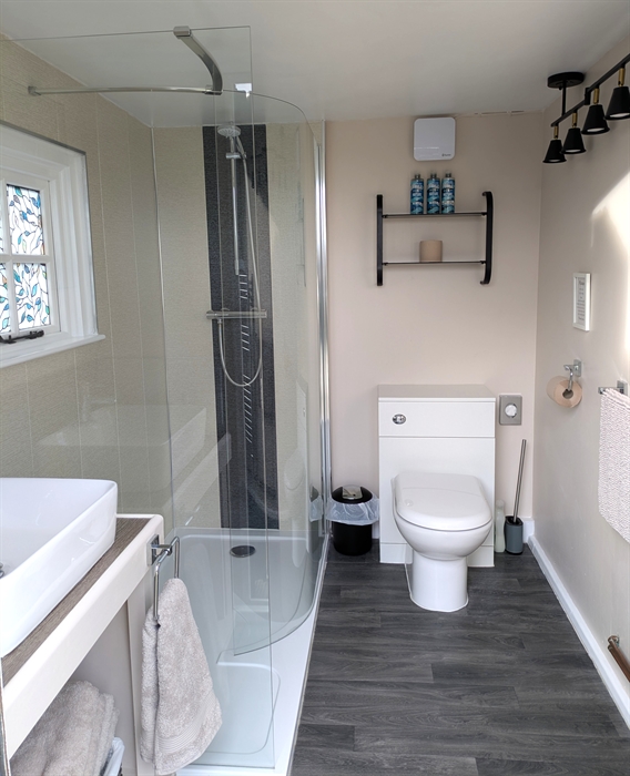 Glass shower cubicle, with cream and charcoal Aquaboard. White toilet and hand basin. Faith in Nature toiletries on black glass shelves. Charcoal viny