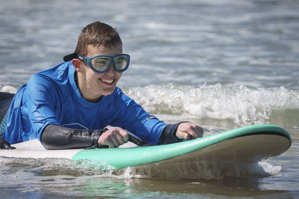 A close up of a young surfer who is disabled, smiling on a wave on a prone surfboard with goggles on