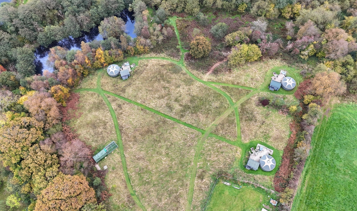 Bird's eye view of 3 yurts in a meadow, surrounded by woodland and with a small lake in top left.