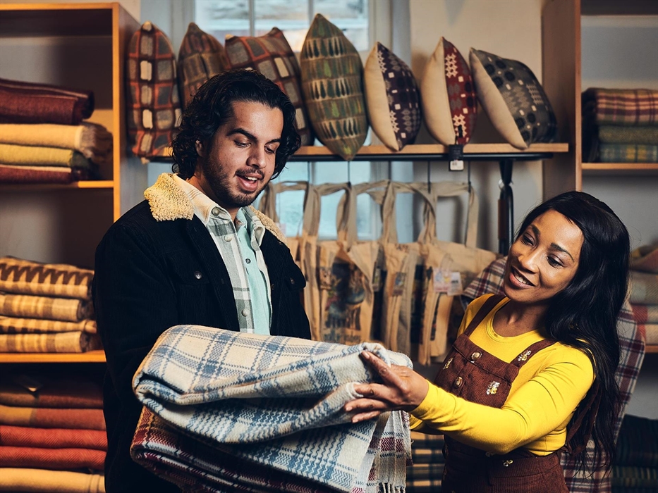 A couple stand in the museum shop, surrounded by blankets, cushions and bags. The man is holding up two woollen blankets as they decide which one to b