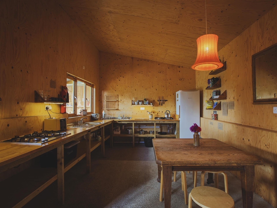 An indoor camp kitchen with a large kitchen space with countertops and hobs. There is a table in the foreground with a light above. A mirror hangs on