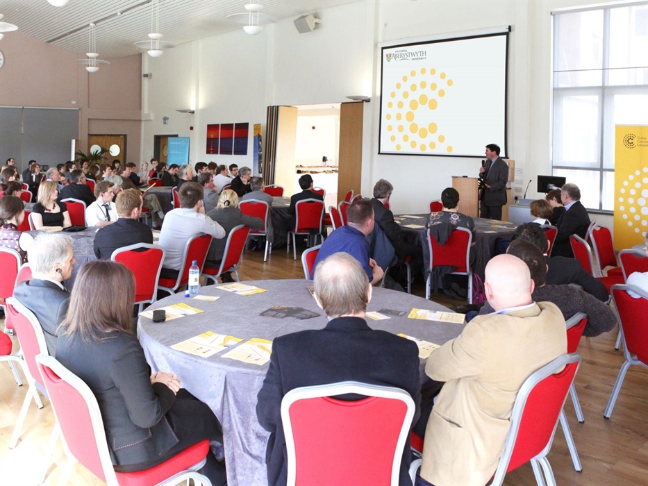 Medrus Mawr Conference Room, delegates sat at round tables viewing a presentation screen.