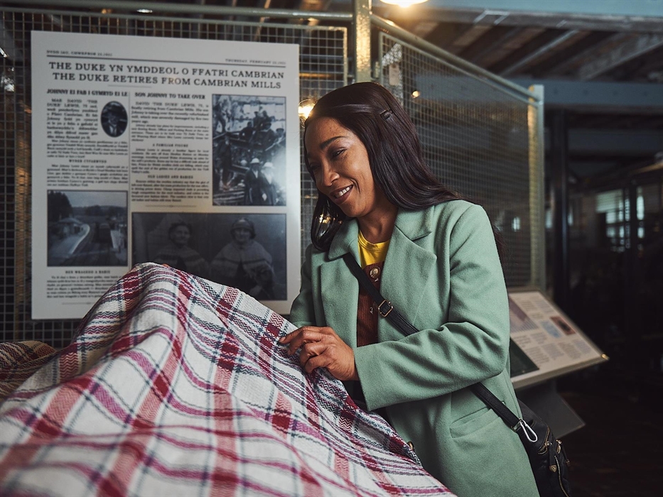 A lady in a green coat stands inside one of the weaving workshops. She is smiling and holding up a woollen blanket to examine it.