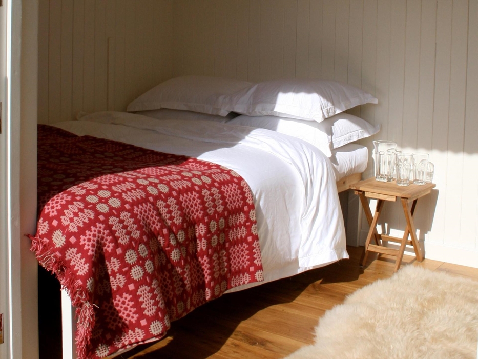 Double bed with the sun shining in with luxurious white cotton bed linen, a red traditional Welsh blanket and a sheepskin rug on the oak floor.