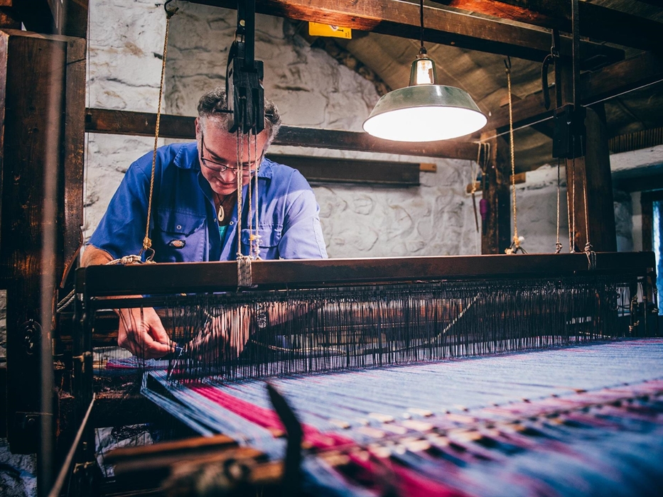 a man in blue overalls works at a traditional loom weaving a blanket