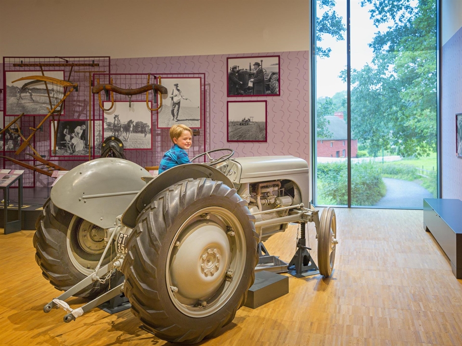 an old grey tractor is inside a museum gallery. A young boy with blond hair and a blue shirt sits on the tractor, as if driving it. There is a view ou