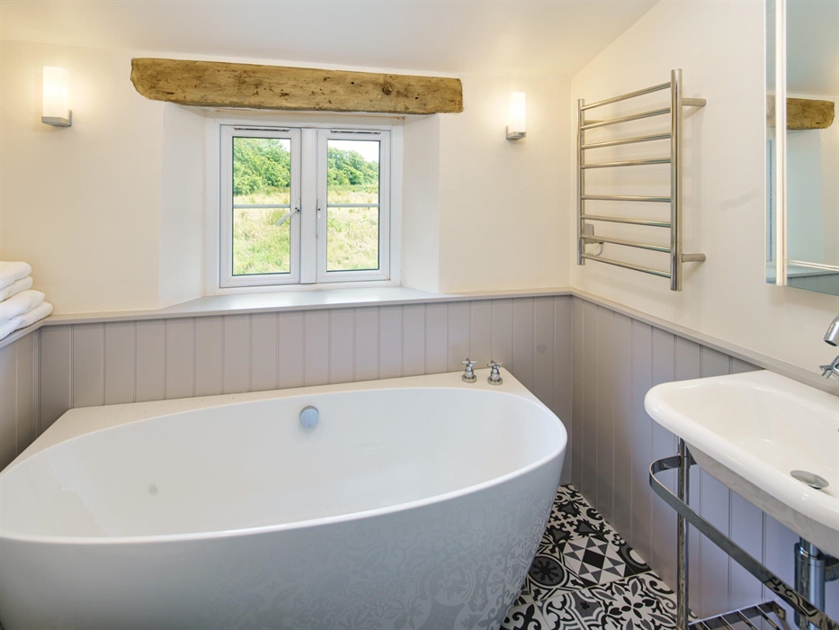 This bathroom has a free standing bath on black and white tiled floor. There is grey tongue and groove panelling, a basin with metal frame, towel rail