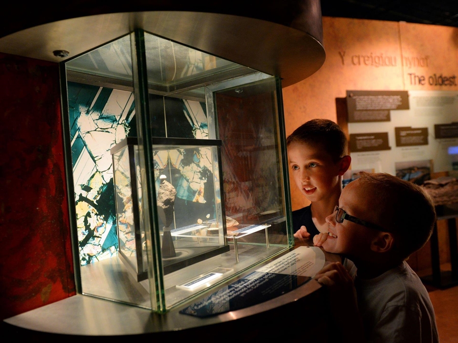 Two young boys are looking at a glass display case in the museum. In the case is a piece of rock, approximately 12 cm tall. The rock was collected by
