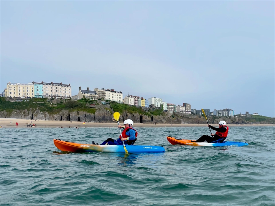 Two adults paddling sit on top kayaks on the sea with Tenby South Beach in the background