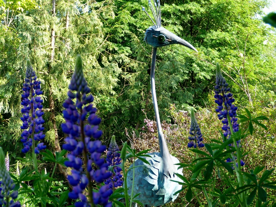 Sculpture of a fantasy peacock made of verdigris copper and glass. Blue lupins in the foreground