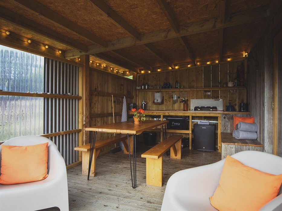 A camp kitchen with festoon lighting above, a countertop with shelving above with glasses and mugs on. A fridge is below the counter and in the foregr