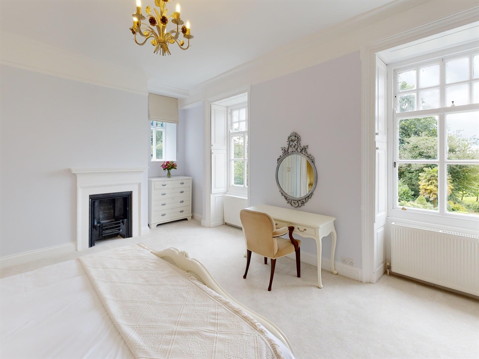Bright and spacious bedroom with a vintage writing desk, ornate mirror, and large sash windows overlooking lush gardens at Plas Cilybebyll.