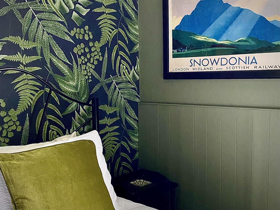 A bedroom at Crafnant House with a green wall, a leaf patterned wallpaper, a green cushion on a white bed and a poster saying "SNOWDONIA"