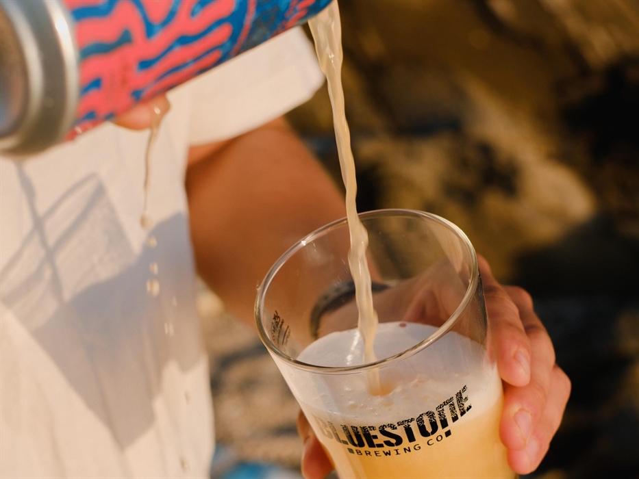 A can of beer is being poured into a glass on a sunny day, the glass has the Bluestone Brewing Co logo printed on the side