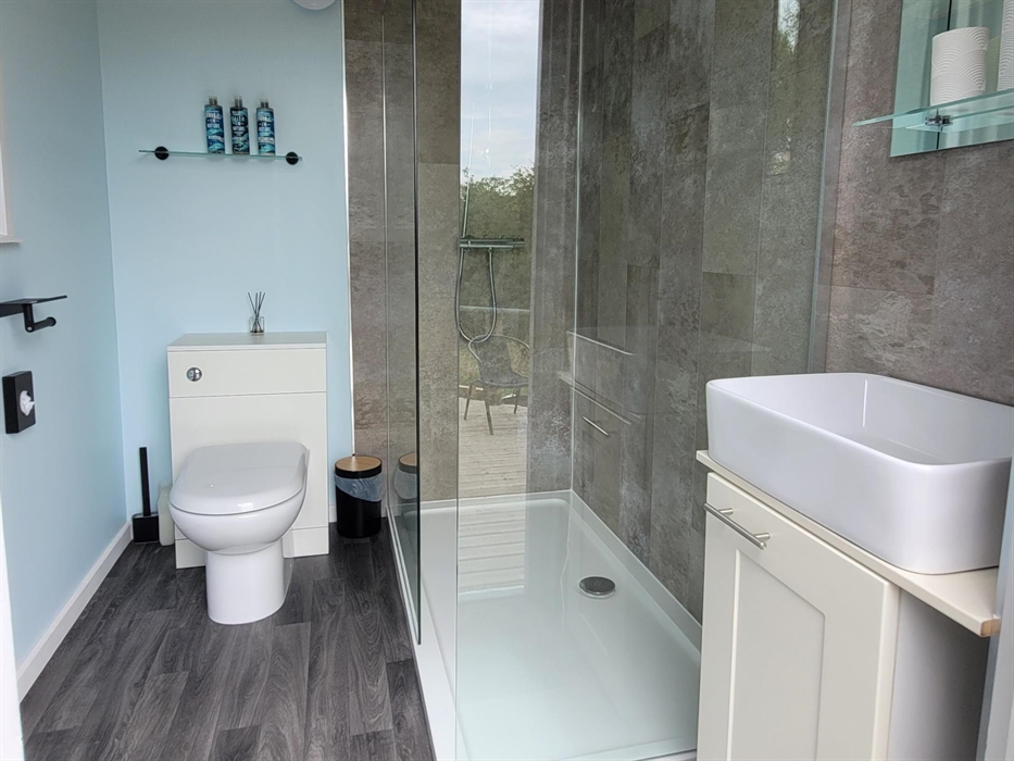 A glass shower enclosure, with brown marble Aquaboard, white toilet and handbasin. The glass shelf above the toilet holds Faith in Nature toiletries.