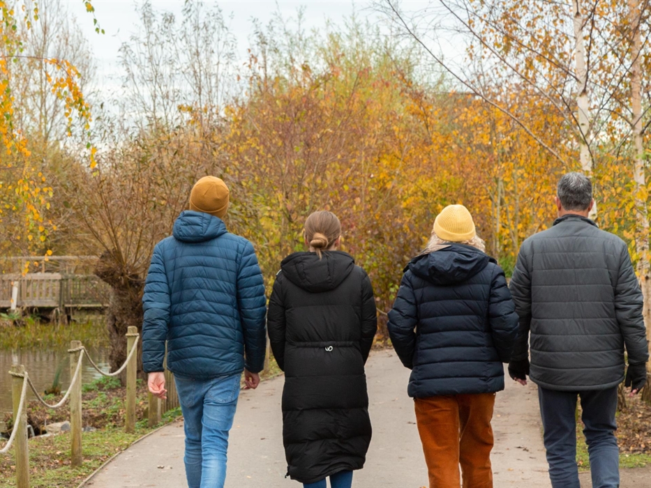 A group walk through an autumnal scene alongside a pond. Yellow and orange leaves against the grey autumn sky.