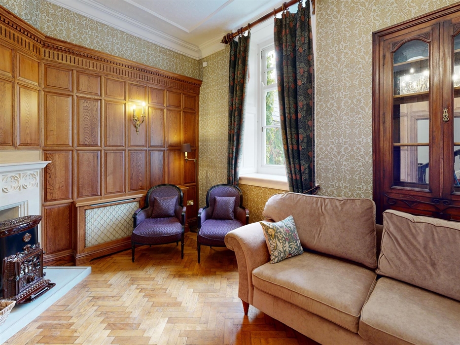 Traditional manor house sitting room with oak paneling, vintage fireplace, patterned wallpaper, curiosity cabinet and plush seating including a velvet