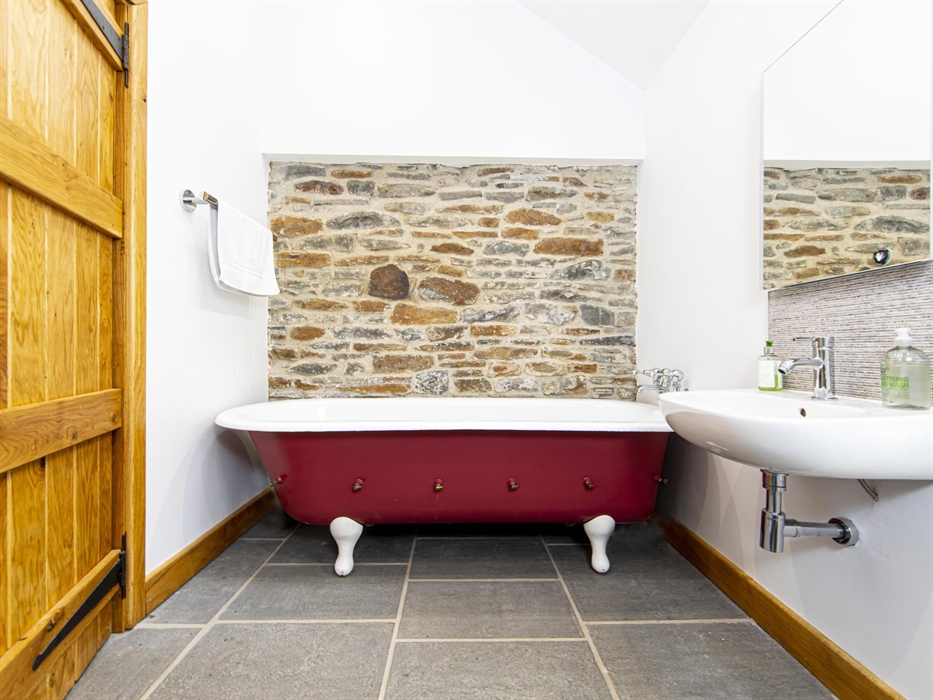 A bathroom with a red roll-top bathtub, white sink, slate floor tiles, and a rustic exposed stone wall, with a wooden door and modern fittings