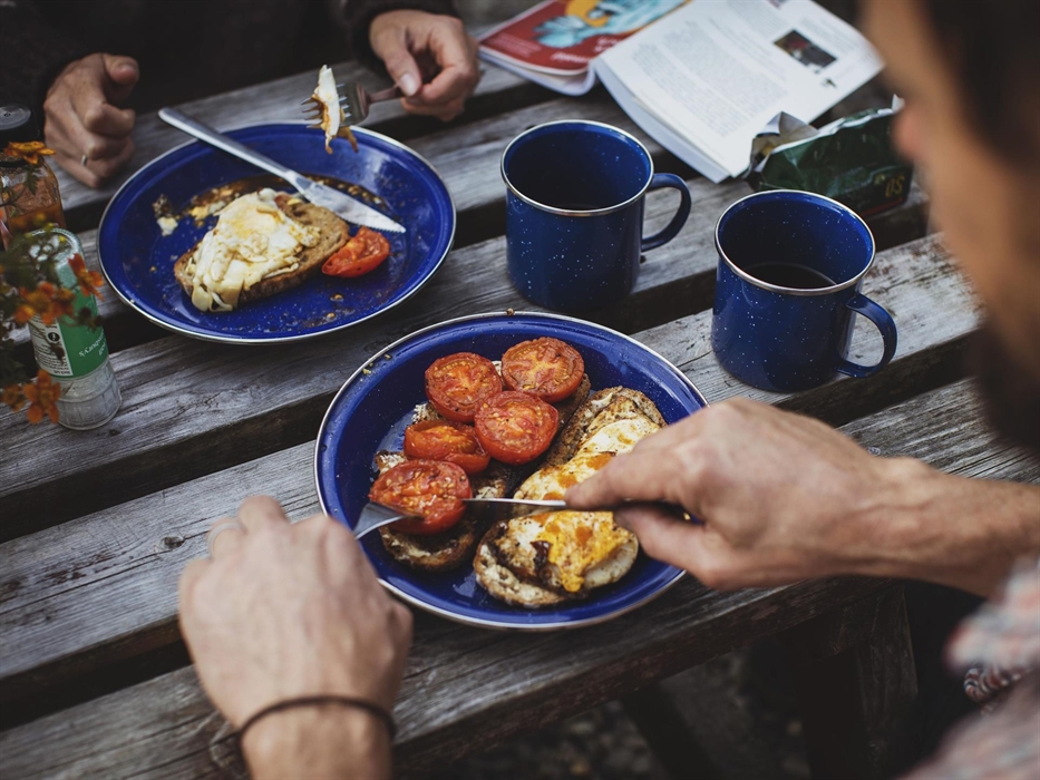 Image taken from above of a couple eating breakfast outdoors. Two plates of fried eggs and tomatoes on toast with cups of hot coffee are visible.
