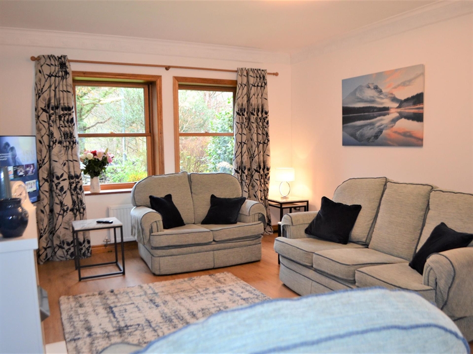 Living room with light blue 3 piece suit, wooden flooring and large television and two wooden windows