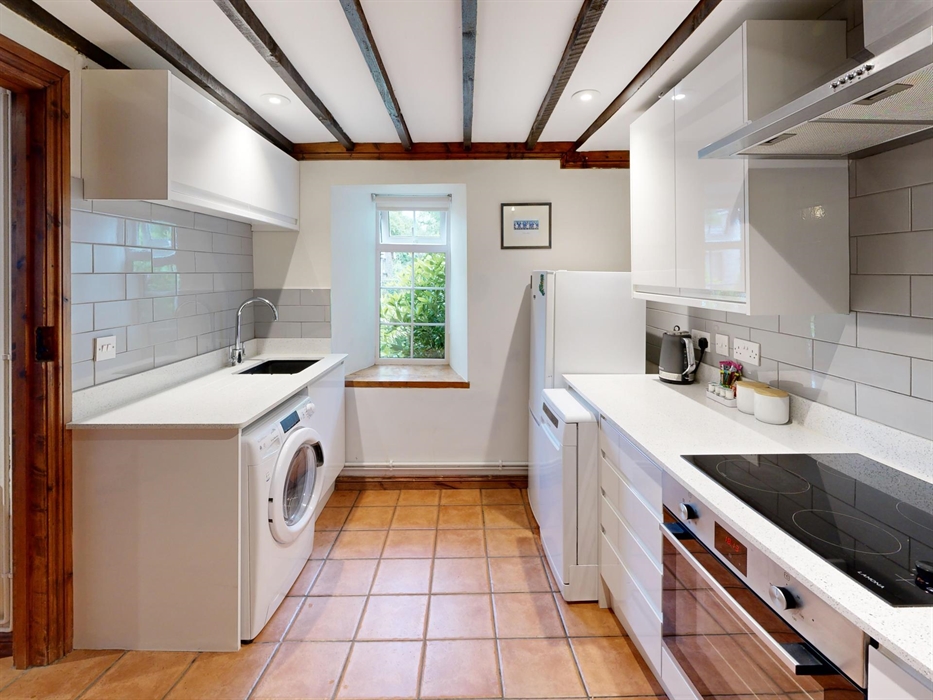 A fully-equipped kitchen with white cabinets, grey tile backsplash, terracotta floor, and a window overlooking greenery.