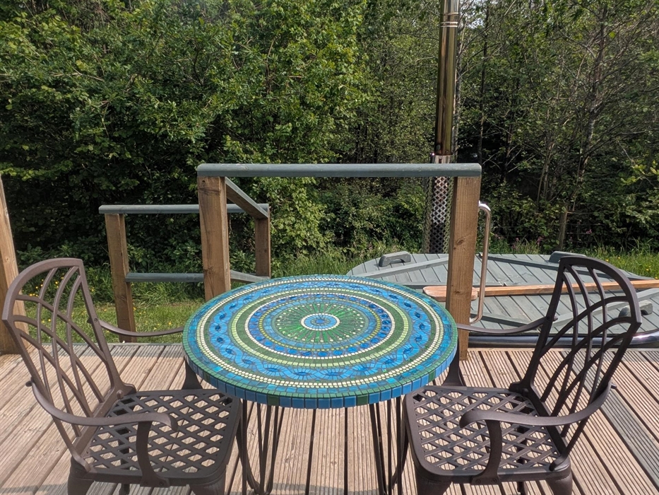 Handmade mosaic table in blue and green with metal dining armchairs on a wood deck with the hot tub, trees and lake in the background.