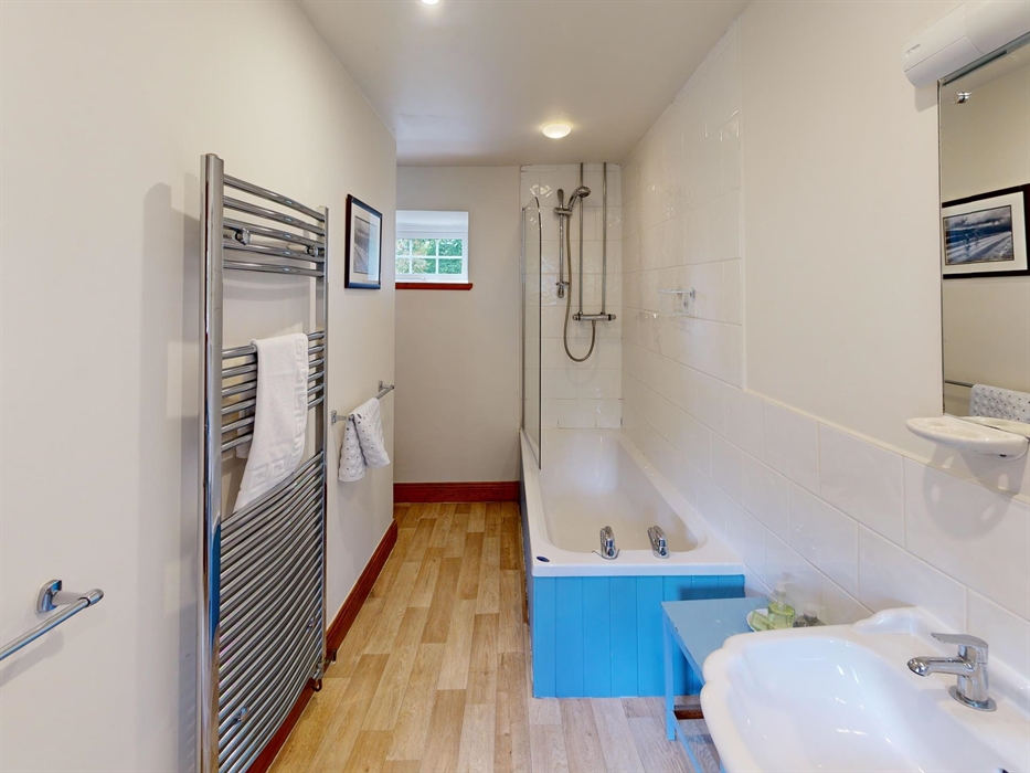 A bright and practical family bathroom featuring a full-sized bath with overhead shower, wooden flooring, a large wall mirror, and chrome towel rails.