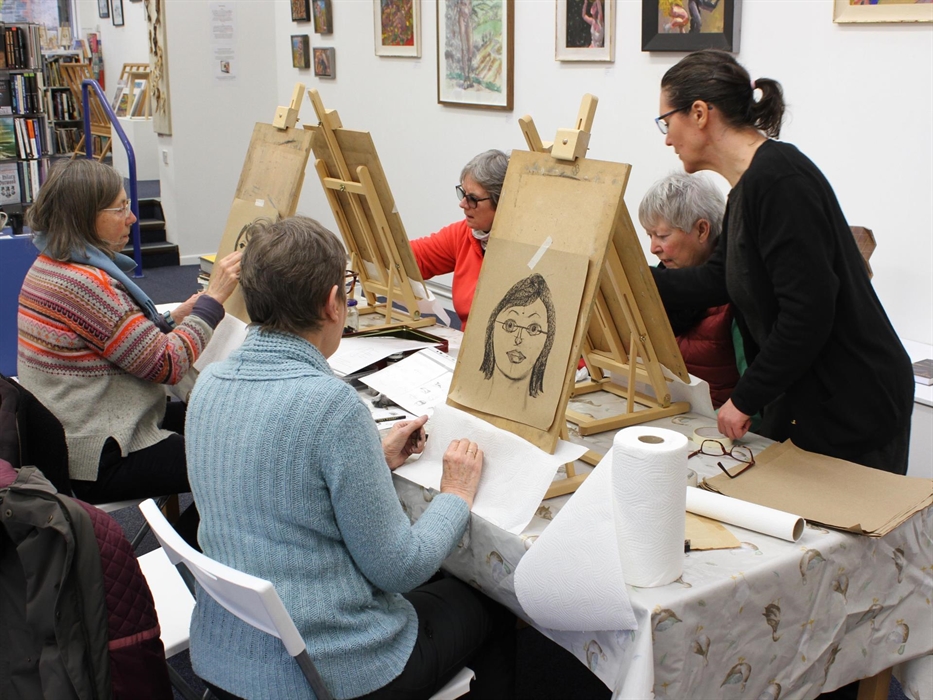 5 figures around a table with 4 seated at wooden easels with drawing boards showing hand drawn portraits. 1 figure is standing. Table is in a gallery
