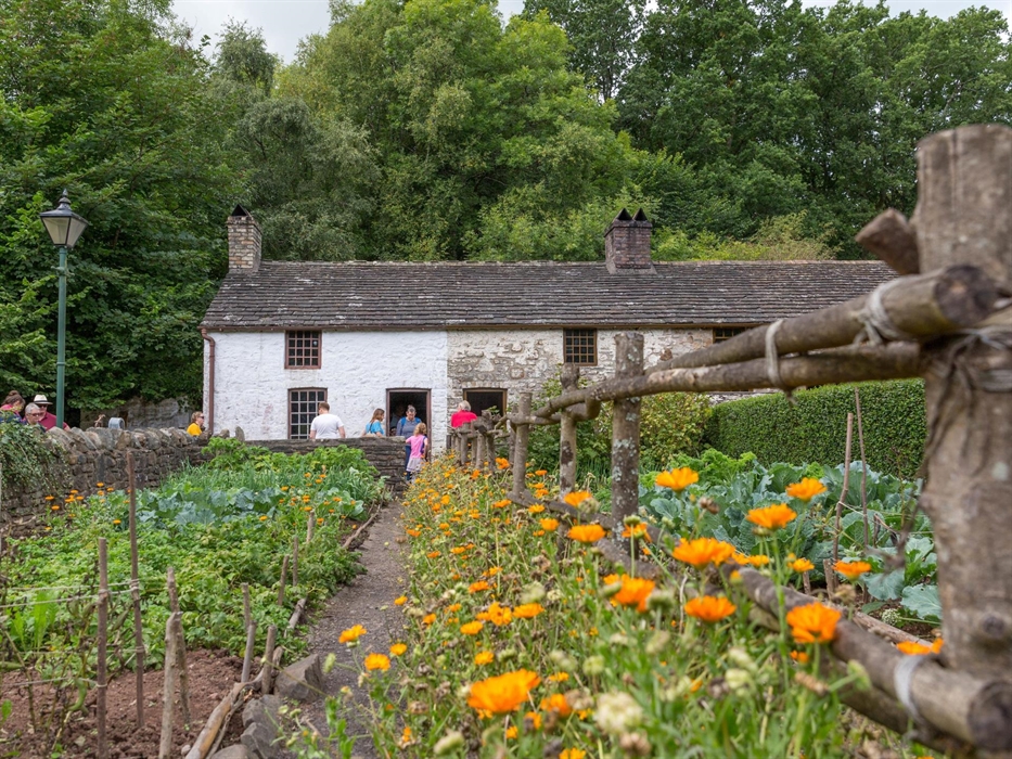 Image shows a short row of 3 small cottages. They have grey tiled rooves and brick chimneys. in the foreground, you can see the cottage garden, plante