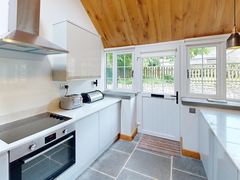 A bright, contemporary kitchen featuring white cabinets, quartz countertops, an electric hob, and a door opening onto a garden with stone walls and fe