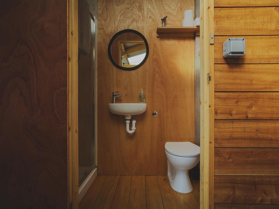 Looking in to a bathroom made of plywood.  There is a warm glow to the image. A shower is seen to the right and in the centre is a basin with mirror a