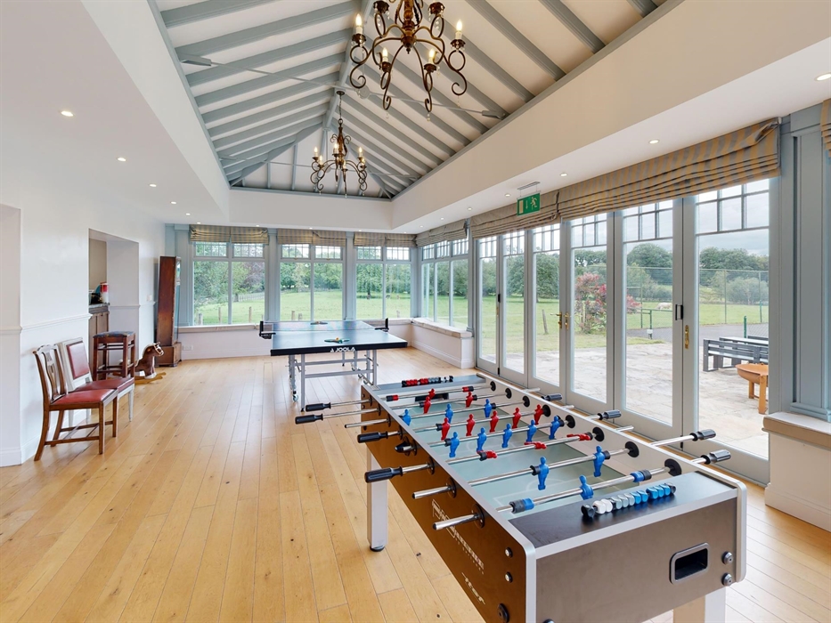 Bright and airy games room with vaulted ceiling, table tennis, and foosball, surrounded by large windows and doors leading to a terrace and tennis cou