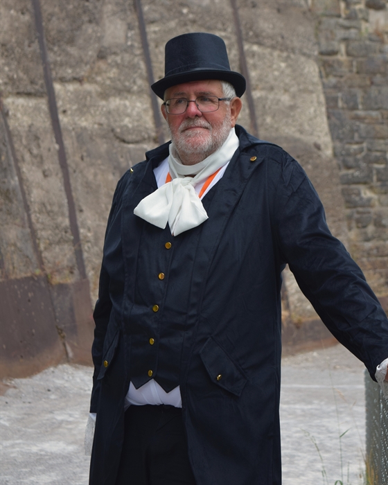 A tour guide dressed up as an 19th Century industrial factory owner standing leaning against a barrier.