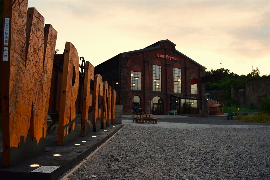Wooden letters spelling Wrecsam in front of a renovated industrial machine shop-turned-coffeehouse at sunset